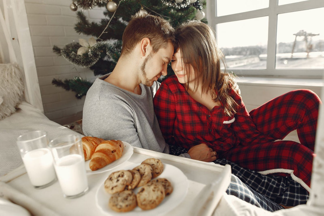 Ein Paar sitzt neben einem Weihnachtsbaum; eine Person trägt einen rot karierten Pyjama.